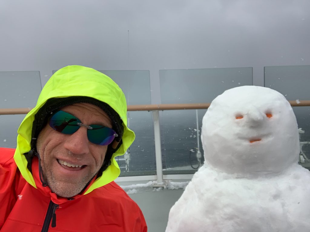 Photo of man in bright orange jacket with yellow hood, sitting next to a snowman on the bow of a ship.