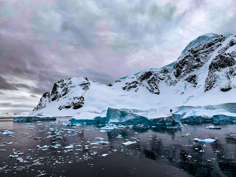 Blue icebergs floating in front of a rocky mountain with snow. Clouds overhead, and reflecting in the water.