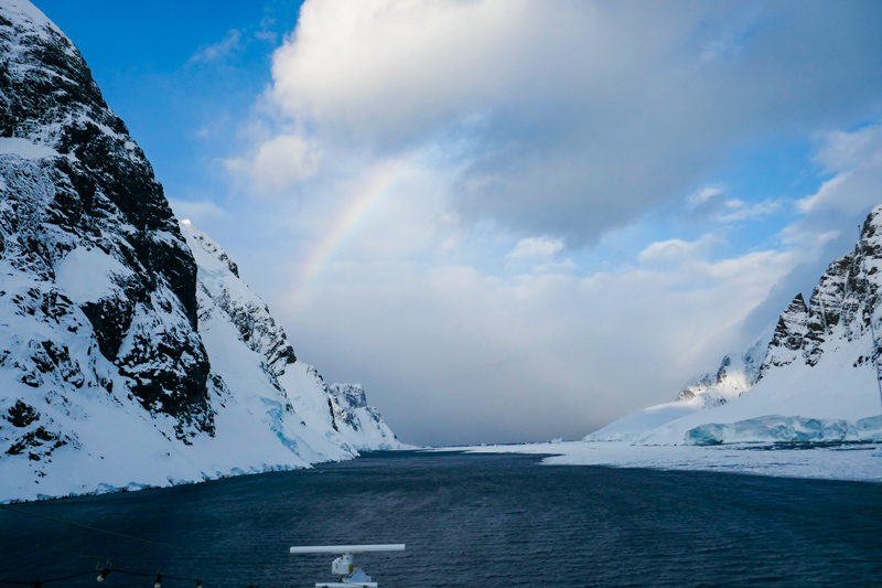 Clouds and a rainbow between two jagged hills straddling a channel with water.