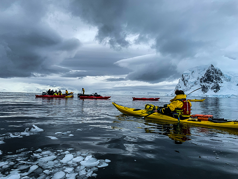 Colorful kayaks and kayakers in water with floating bits of ice, and dark clouds overhead.