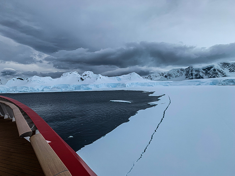 Bow of a ship with red trim cracking a giant ice sheet.