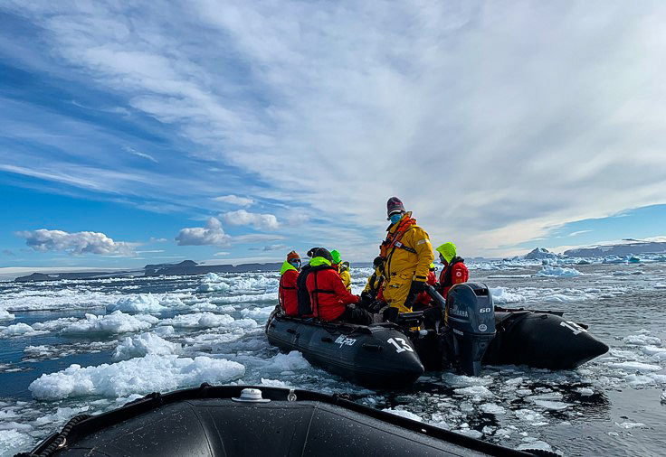 An expedition team, wearing bright orange and yellow jackets, on an inflatable Zodiac boat cruising through water with chunks of ice