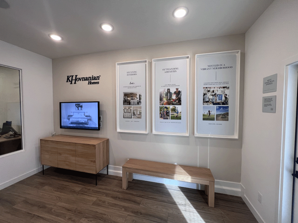 Small room with wood floor, a modern wood credenza and four-legged bench.  On the wall are a touchscreen monitor, with K. Hovnanian Homes logo above and 3 large white framed poster on the right.