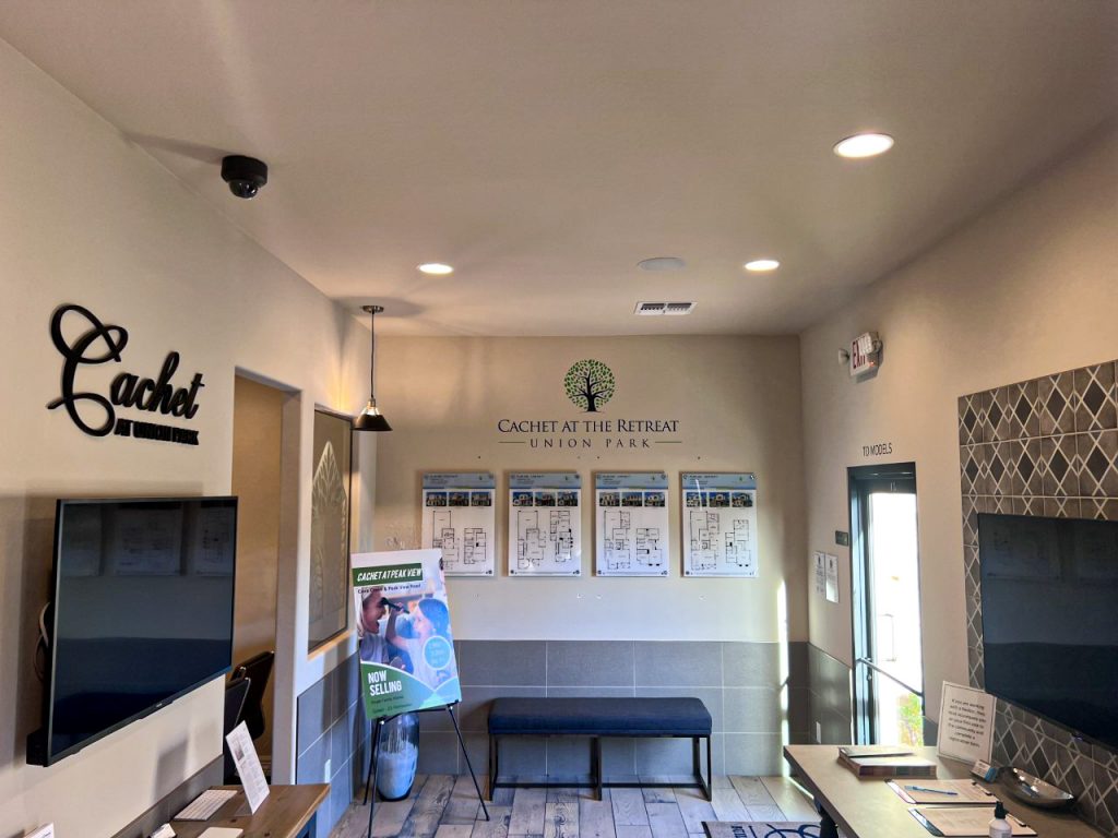 A small sales center (model home center) installation for Arizona builder Cachet Homes. Cachet Logo with monitor on the left wall.  Union Park logo with floor plan posters, and a blue bench on the facing wall. Touch screen monitor for an interactive kiosk on the right wall with a table below holding print materials.