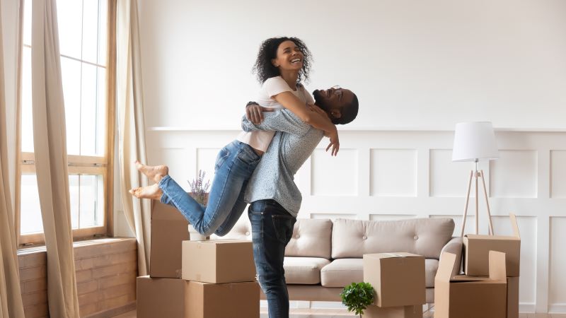 Happy Homeowners, man and woman hugging. Man lifts woman into the air. Boxes scattered around room ready to unpack.