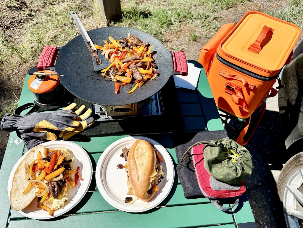 An assortment of cookware and food om a pistachio-colored camp table next to an orange-colored ice chest.