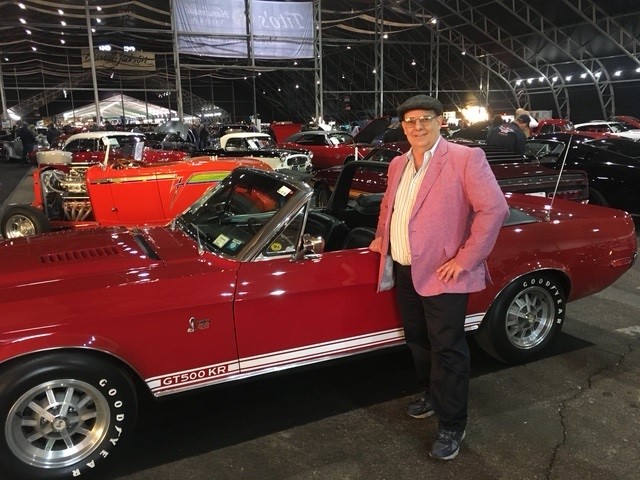 Man sharply dressed in pink blazer and driving cap next to bright red antique convertible sportscar.